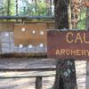 A sign that says, "Caution: Archery Range" sits in front of a small wall filled with hay that has hay bales in front of it.