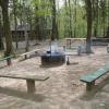 View 2 of a circular fire pit surrounded by benches and trees with a camp pavilion in the background.