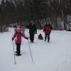 Two adults walk as several children snowshoe through the woods. One adult pulls a child in a sled.