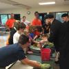 Two men in black chef suits talk to several children next to a long table bearing fruits and vegetables.