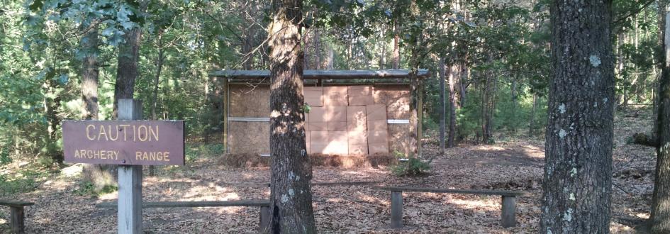 A sign that says "Caution: Archery Range" sits in front of two benches and a small wooden wall.