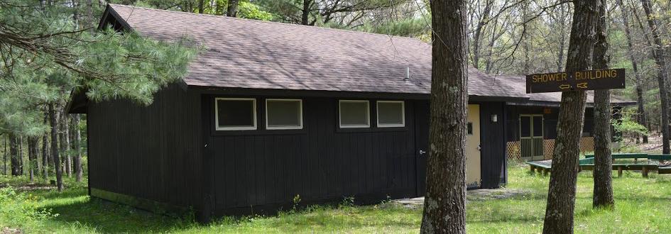 A brown shower building in the foreground and screened-in Jack Pine Pavilion in the background, surrounded by trees.