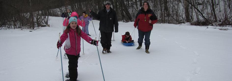 Two adults walk as several children snowshoe through the woods. One adult pulls a child in a sled.